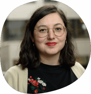 Franziska trägt eine runde Brille, geblümte Bluse und roten Lippenstift. Sie hat schulterlanges braunes Haar