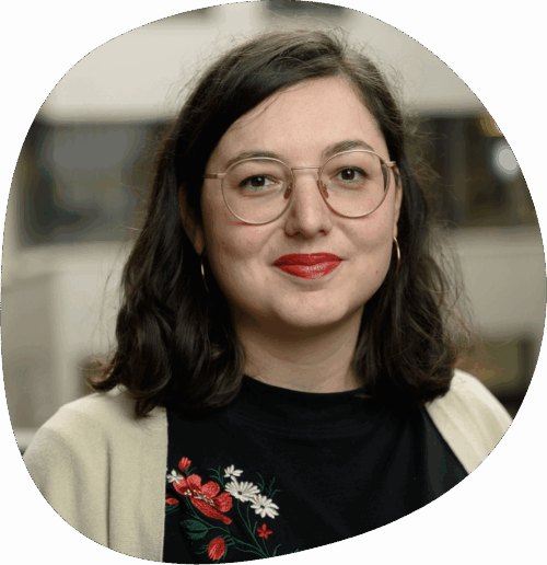Franziska trägt eine runde Brille, geblümte Bluse und roten Lippenstift. Sie hat schulterlanges braunes Haar