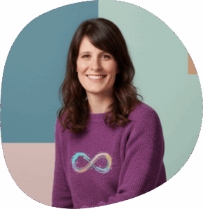 Sarah trägt schulterlange braune Haare und einen lila Pullover mit dem Neurodiversitäts-Symbol darauf: eine auf der Seite liegende Acht in Regenbogenfarben