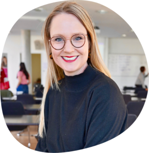 Sarah hat lange blonde Haare und eine Brille. Sie trägt einen schwarzen Rollkragenpullover und roten Lippenstift.