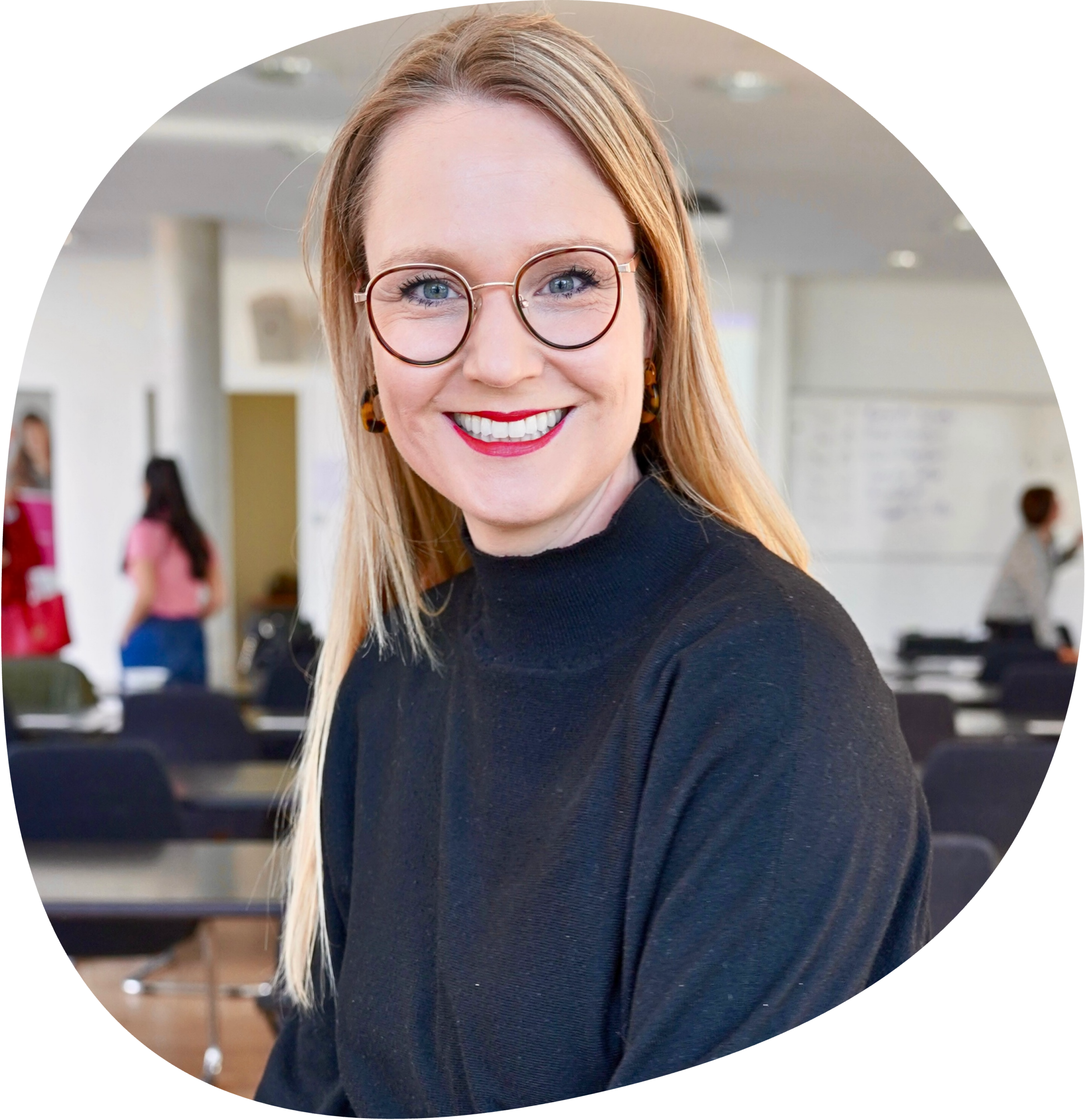 Sarah hat lange blonde Haare und eine Brille. Sie trägt einen schwarzen Rollkragenpullover und roten Lippenstift.