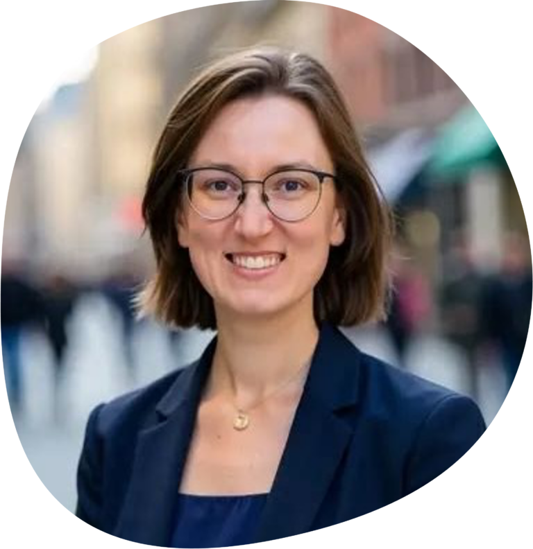Saskia trägt kinnlange braune Haare, eine Brille und einen dunkelblauen Blazer