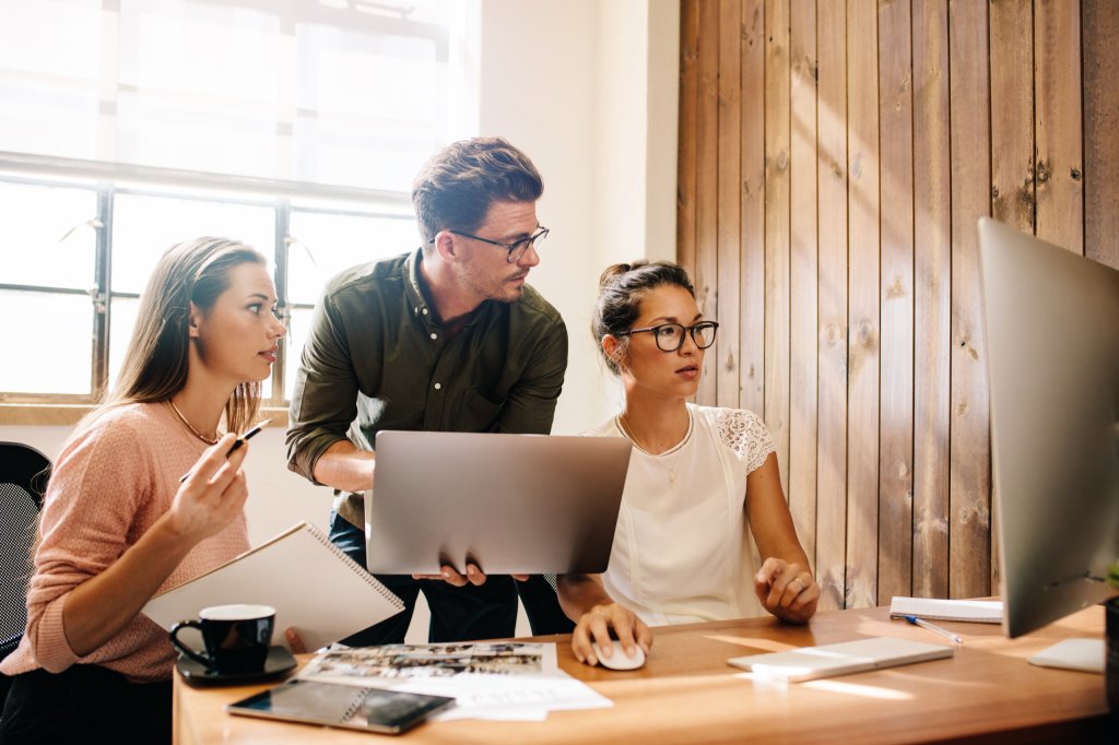 Drei Personen schauen gemeinsam auf einen Laptop und einen Monitor