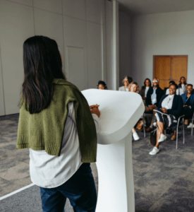 Eine Frau steht am Redepult und hält eine Keynote vor einer Gruppe Menschen, die auf Stühlen vor ihr sitzen