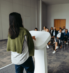 Eine Frau steht am Redepult und hält eine Keynote vor einer Gruppe Menschen, die auf Stühlen vor ihr sitzen