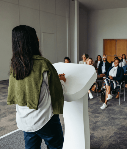 Eine Frau steht am Redepult und hält eine Keynote vor einer Gruppe Menschen, die auf Stühlen vor ihr sitzen