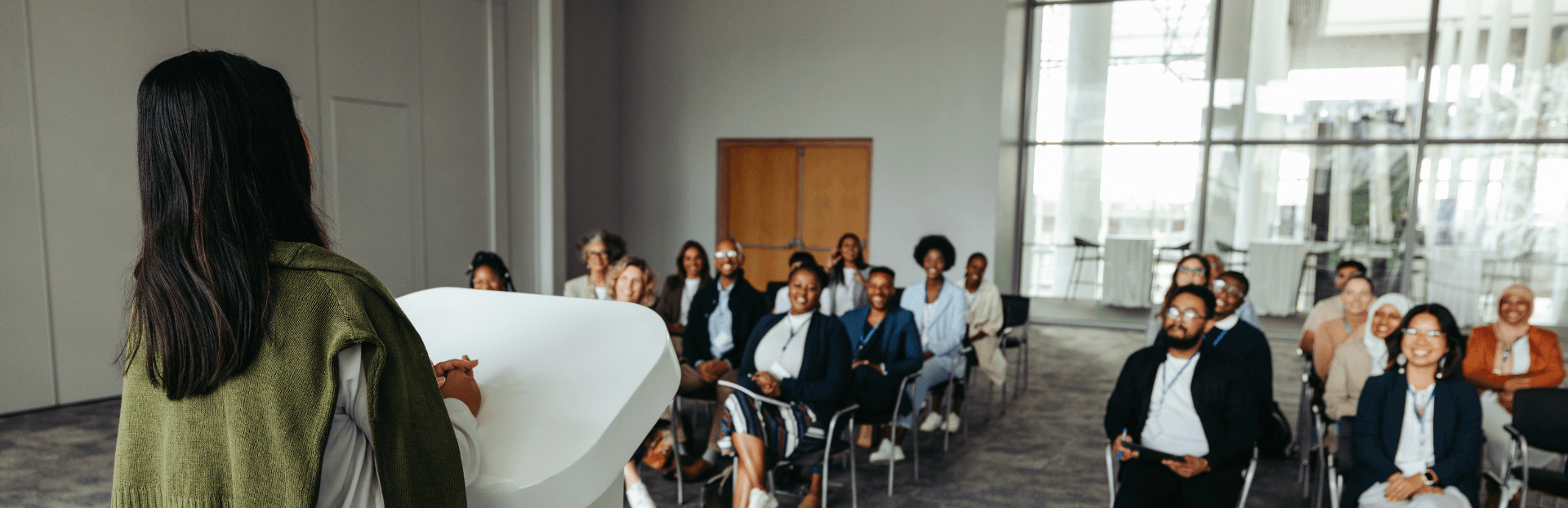 Eine Frau steht am Redepult und hält eine Keynote vor einer Gruppe Menschen, die auf Stühlen vor ihr sitzen